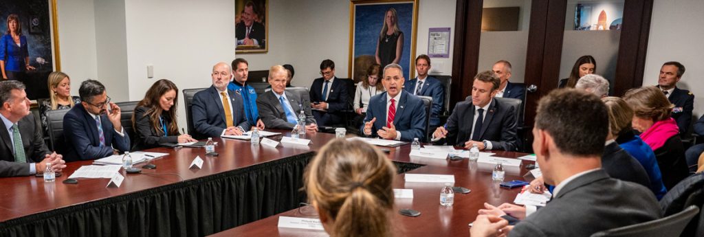 Executive boardroom table full of people, including Deputy Secretary of Commerce Don Graves (speaking/gesturing at center), French President Emmanuel Macron (right of Graves), NASA Administrator Bill Nelson (left of Graves), NOAA Administrator Rick Spinrad (left of Nelson), and National Space Council Executive Secretary Chirag Parikh (2nd from left)