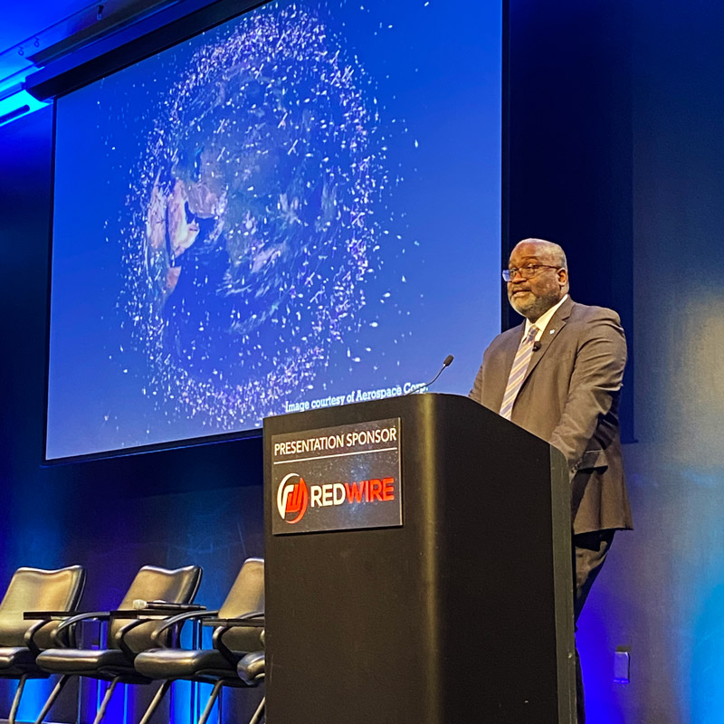 Dr. Michael Morgan at podium with image of space traffic around Earth in the background