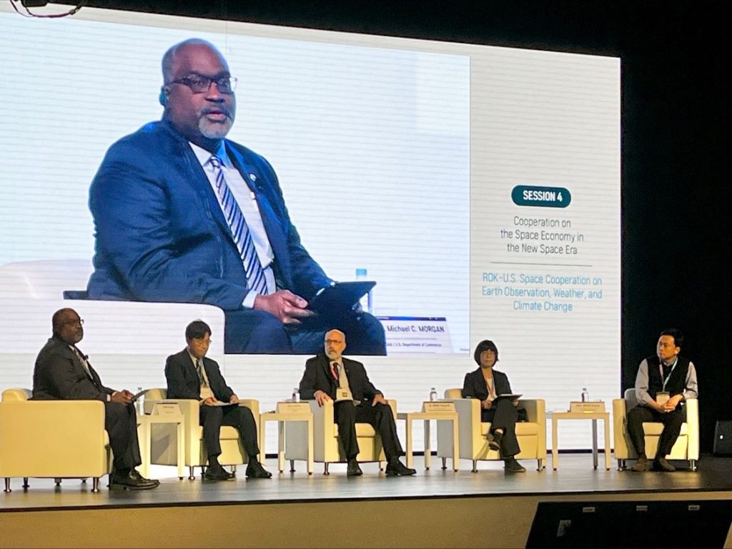 Michael Morgan appears on a large screen as he addresses four other seated panelists on stage