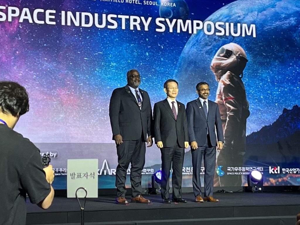 NOAA Assistant Secretary Michael Morgan, MSIT Minister Jong-Ho Lee, and National Space Council Executive Secretary Chirag Parikh pose before a graphic backdrop reading “Space Industry Symposium” and featuring a standing astronaut looking up into space