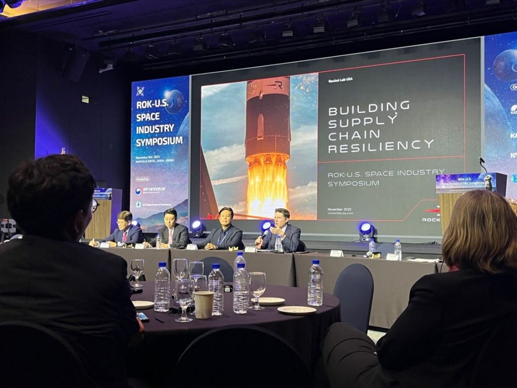 Seated panelists speak before an audience with a backdrop of a rocket reading “Building Supply Chain Resiliency”