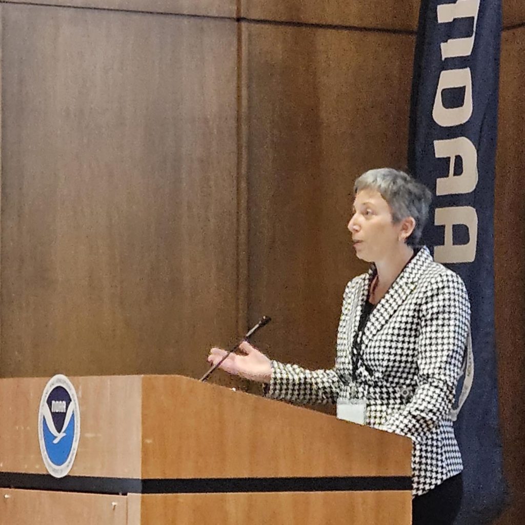 Janice Starzyk speaks from a podium with a NOAA logo in front and a NOAA banner behind her
