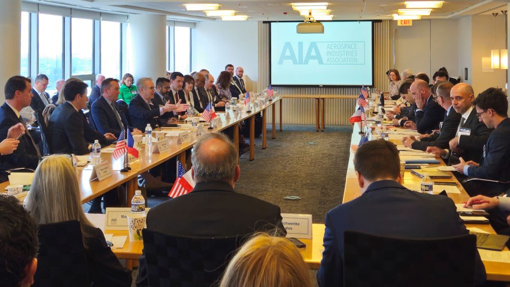 Conference table with two delegations facing each other and the AIA logo projected on the screen between them. OSC Director Richard DalBello (left center) is gesturing while speaking.