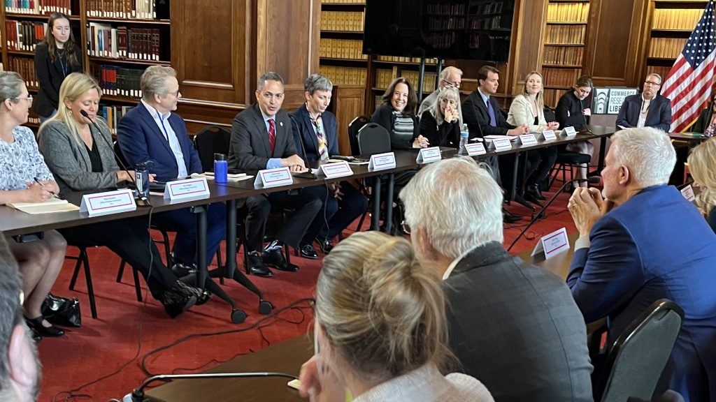 Deputy Secretary of Commerce Don Graves (wearing red tie) speaks from table to a group of people in a library with the U.S. flag on the right