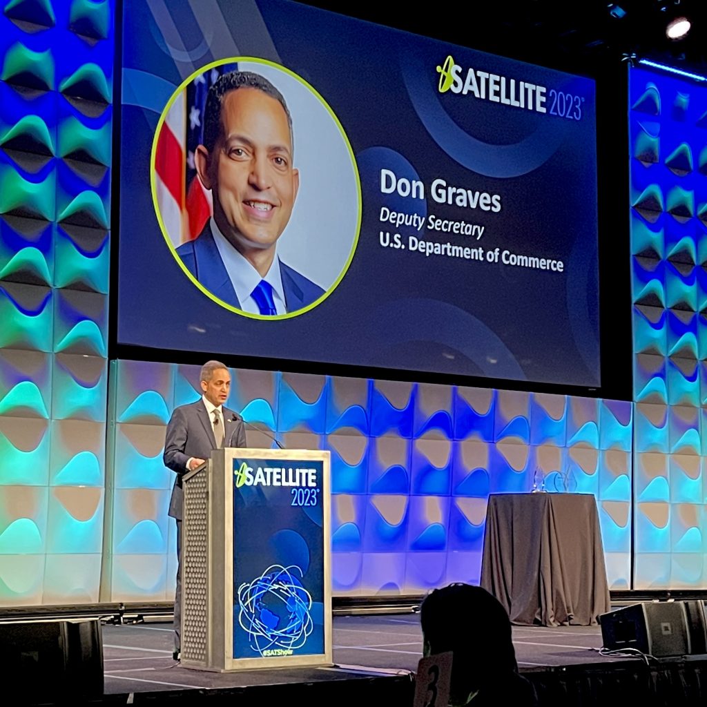 Deputy Secretary Don Graves standing at podium on stage with colorful backdrop and SATELLITE 2023 graphics